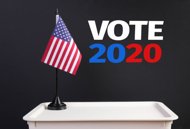 Black background with American flag atop small white desk. Text reads "Vote 2020."